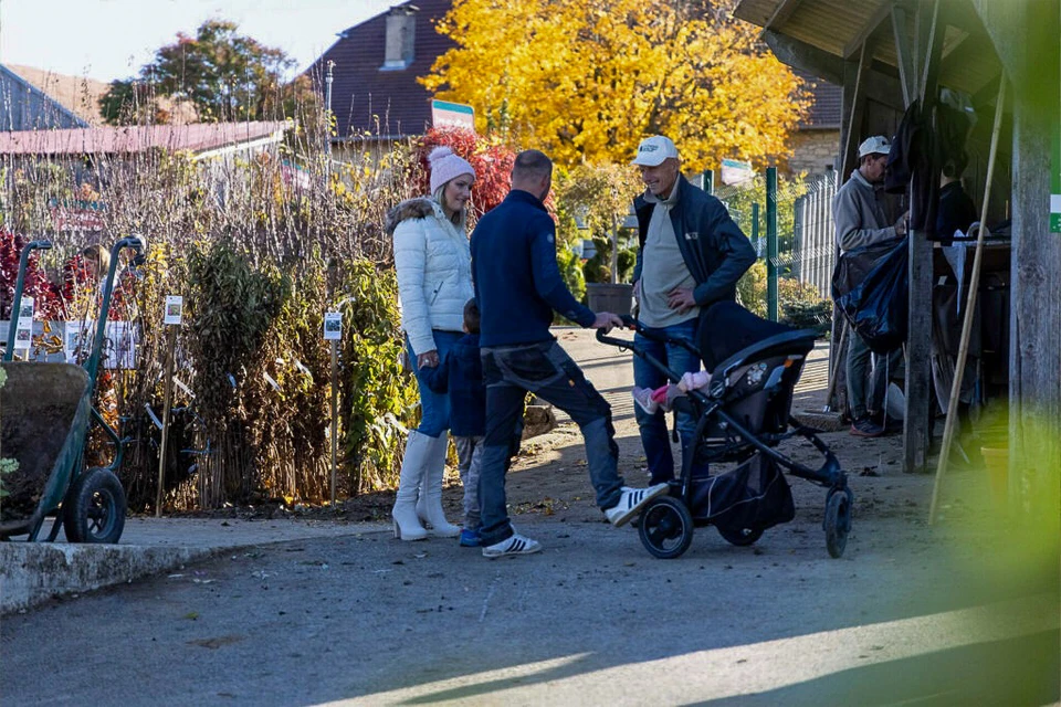 Homme barbu souriant en tablier beige et chemise bleue tenant une caissette en bois remplie de jeunes plants verts devant sa boutique. Devanture avec grandes baies vitrées noires, présentoirs métalliques exposant diverses plantes en pots colorés, chrysanthèmes et plantes aromatiques.
